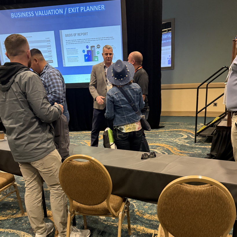 Business professionals discussing business valuation strategies at a conference, with a presentation on exit planning in the background.
