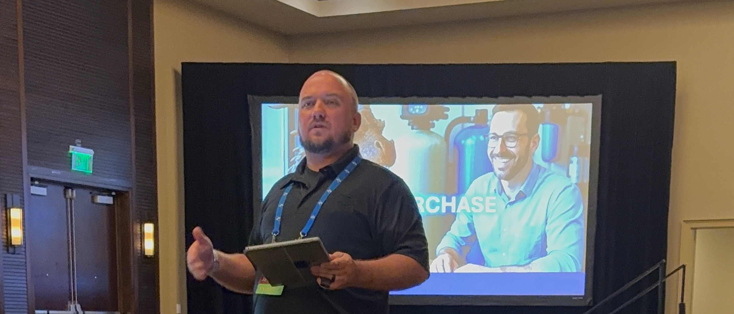 Man presenting at a business seminar with a screen behind him displaying a smiling individual, emphasizing business purchase strategies and growth opportunities.