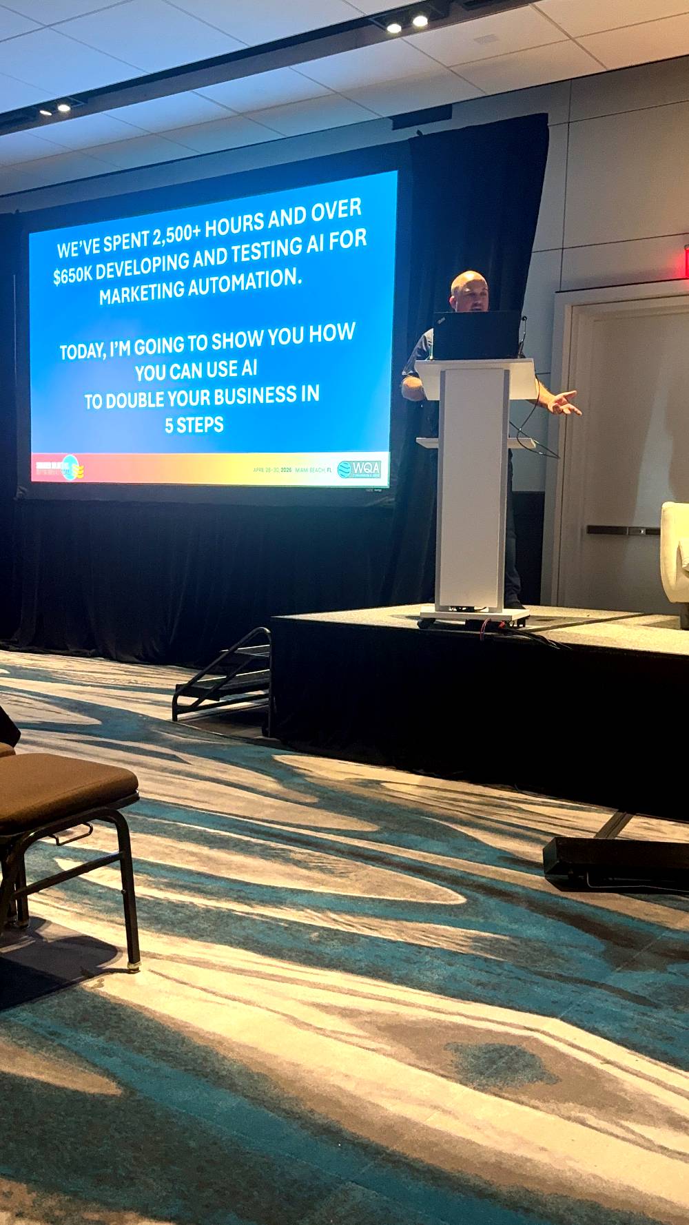 Speaker presenting on stage about using AI for marketing automation, with a large screen displaying text on doubling business in five steps.