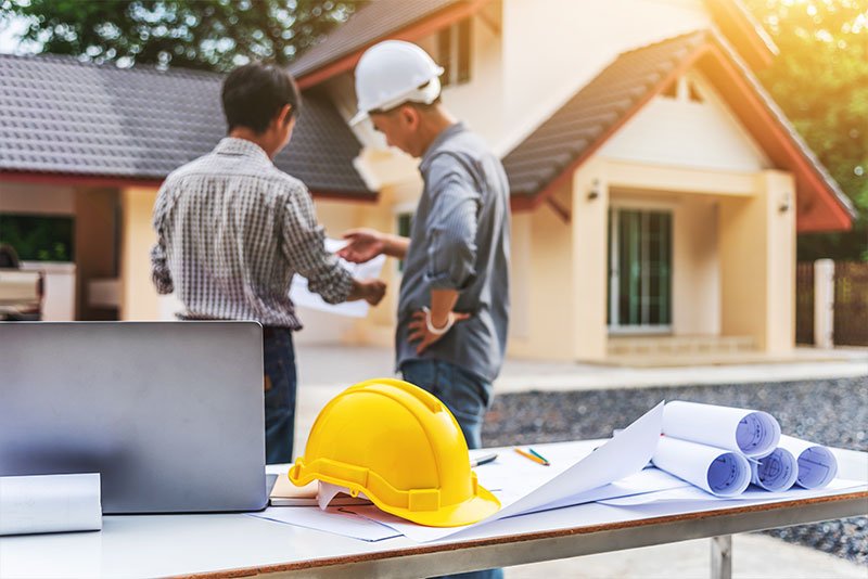 Two men discussing home improvement plans outside a residential property, with a yellow hard hat and blueprints on a table, emphasizing the remodeling industry in the Midwest.