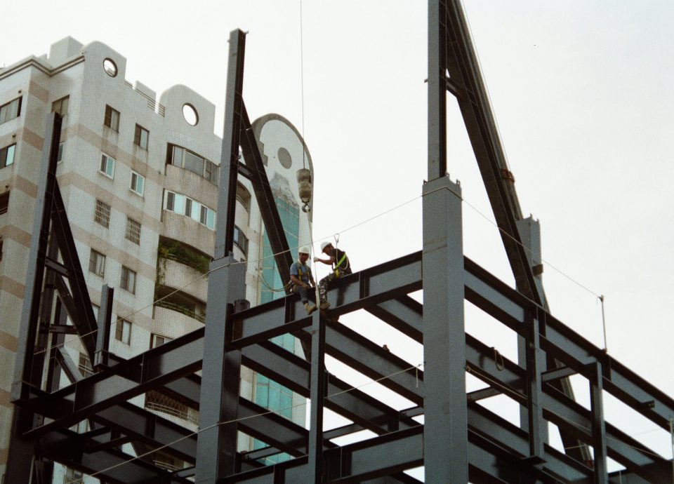 Construction workers assembling steel framework for a building, with a modern structure in the background.