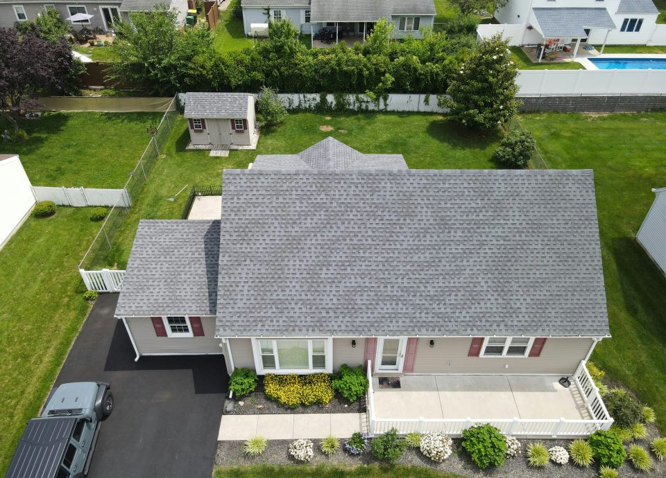 Aerial view of a residential home with a gray roof, landscaped yard featuring flowers and greenery, and a nearby shed, highlighting potential property for sale or investment in real estate.