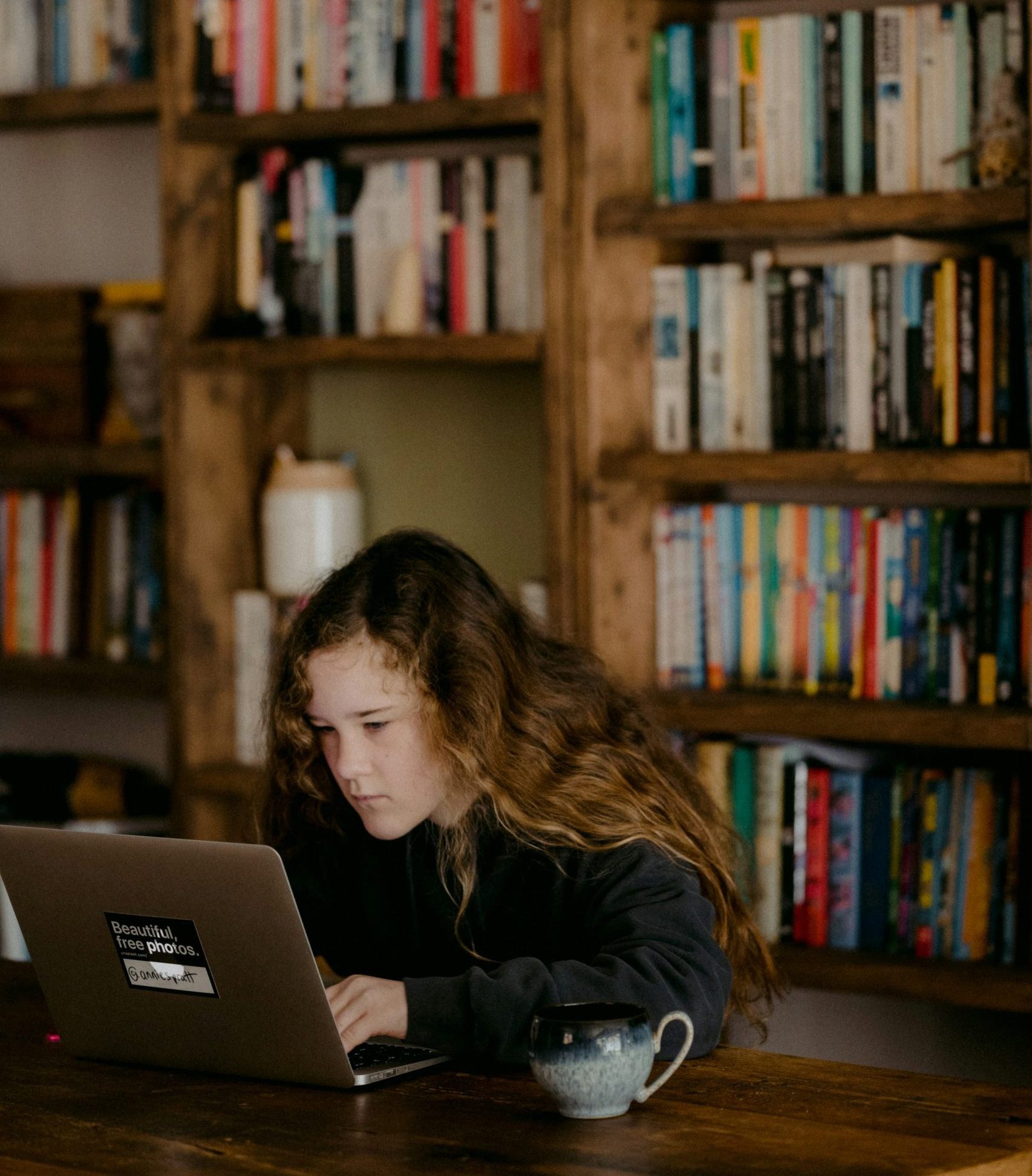 Young person using a laptop at a wooden table with a mug, surrounded by bookshelves, illustrating the educational focus of Captyx's services for education business owners.