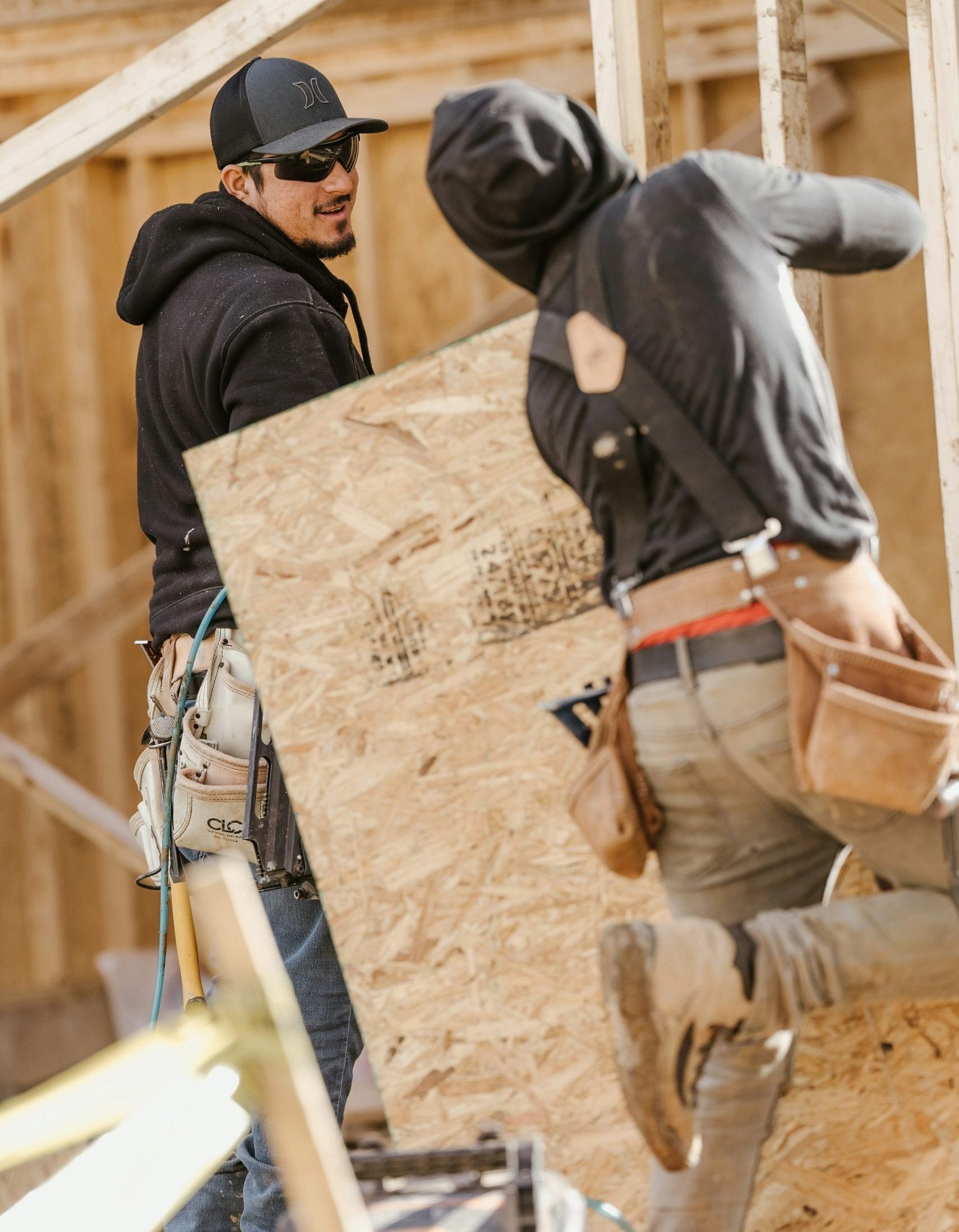 Two construction workers on-site, one smiling while holding a large piece of plywood, showcasing teamwork in general contracting.